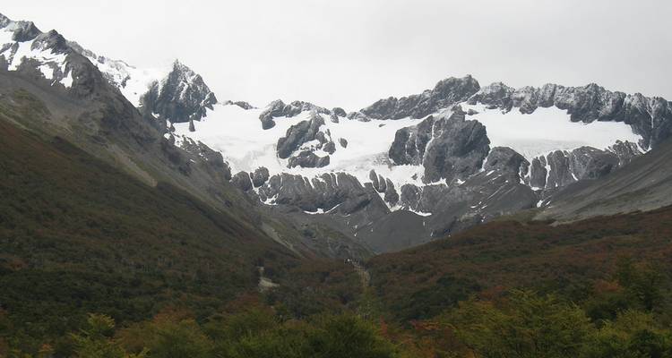 Des pics escarpés et accidentés avec des glaciers suspendus dominent une vallée boisée sous un ciel gris.