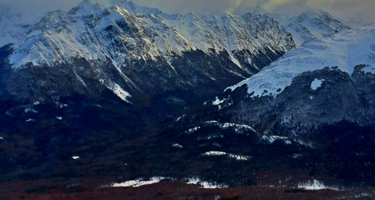 Vista de teleobjetivo de estado de ánimo de picos nevados y accidentados sobre laderas boscosas oscuras.