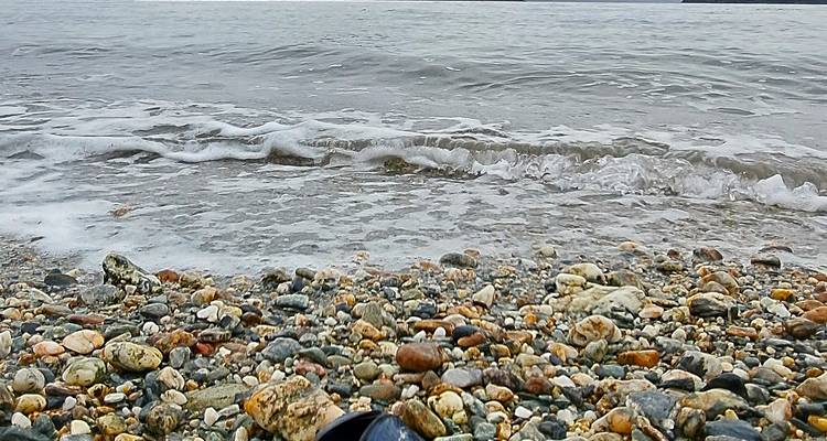 Vista de cerca de guijarros coloridos en una playa mientras las olas suaves lamen la orilla bajo un cielo nublado.