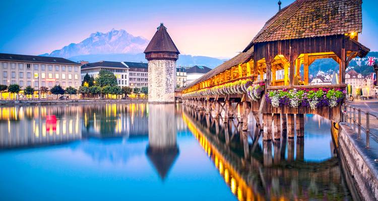 Le pont de la Chapelle et la tour de Lucerne se reflétant dans une eau calme avec un décor montagneux au crépuscule.