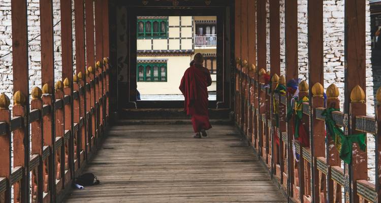 Mönch, der über eine traditionelle Holzbrücke in Bhutan geht, mit architektonischen Details im Hintergrund.