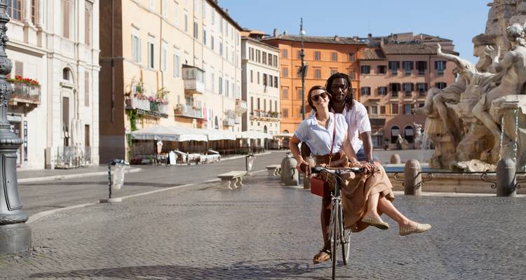 Ein Paar auf einem Fahrrad auf einem malerischen Platz mit historischen Gebäuden und einem Brunnen.