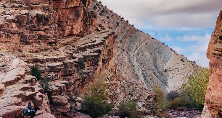 Un excursionista atraviesa un dramático cañón de rocas rojas con acantilados en capas bajo un cielo nublado.