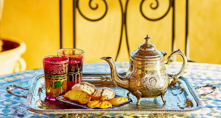 Tetera marroquí tradicional de plata, vasos y pasteles en una bandeja ornamentada sobre una mesa de mosaico.