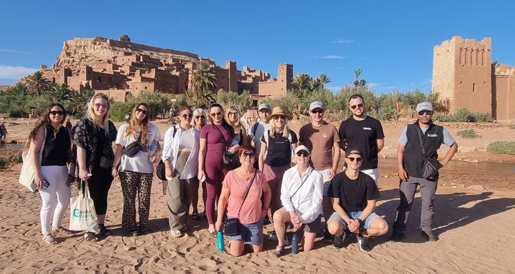 Grupo de turismo posando en la orilla arenosa del río con la famosa kasbah del desierto y sus torres detrás de ellos.
