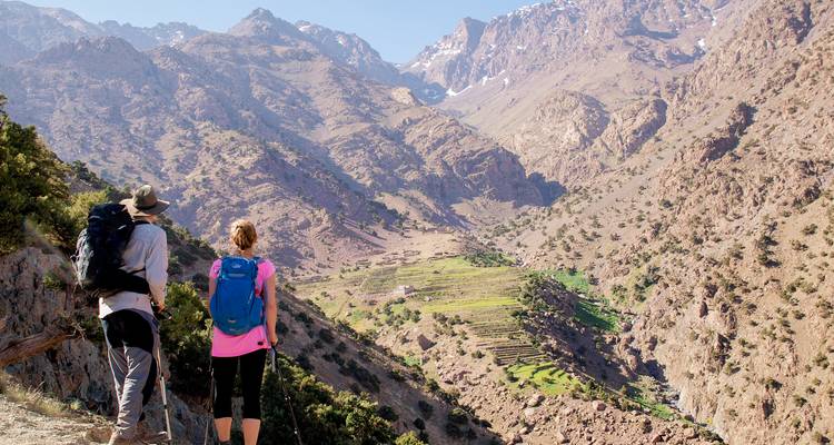 Dos excursionistas hacen una pausa en un sendero de montaña que da vista a un valle verde en terrazas en el Alto Atlas.