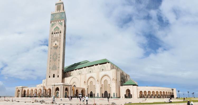 De imposante Hassan II-moskee torent boven een plein uit met kleine bezoekers beneden en een gedeeltelijk bewolkte hemel.