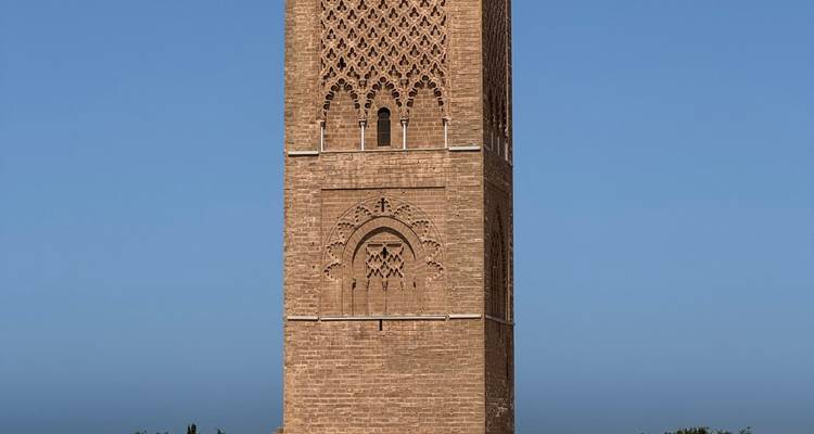 De rode stenen Hassan Tower steekt hoog uit boven de boomtoppen tegen een wolkenloze blauwe lucht.