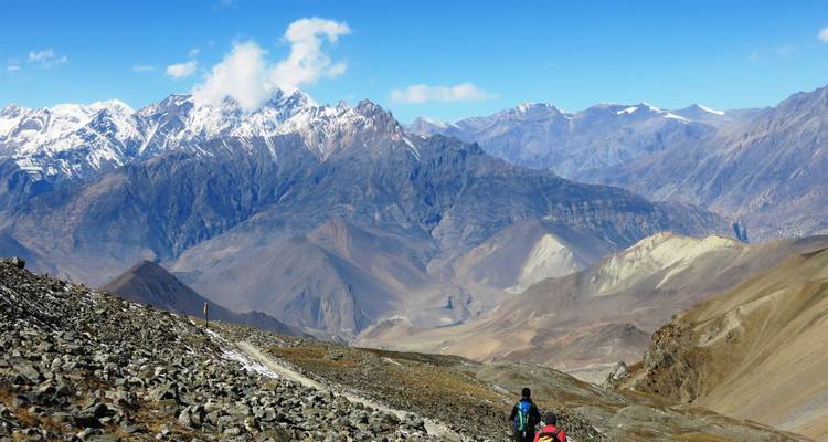 Un dramático paisaje de montaña con un sendero que conduce a dos excursionistas por terreno rocoso.