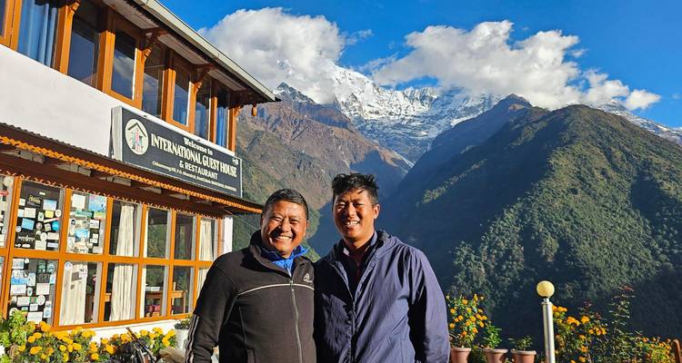 Dos hombres sonrientes están parados afuera de una casa de huéspedes con los nevados picos del Himalaya asomándose detrás.