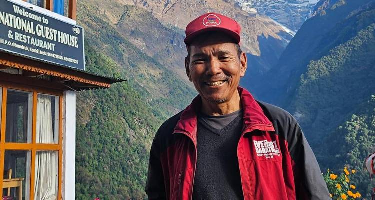 Retrato de un hombre sonriendo con una gorra roja parado frente a un letrero de casa de huéspedes con profundos valles del Himalaya de fondo.