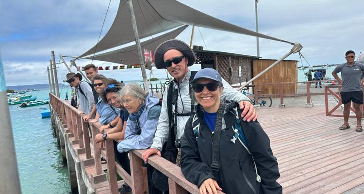 Een groep reizigers leunt tegen een houten pier railing en kijkt uit over helder turquoise water met kleine bootjes onder een gedeeltelijk bewolkte lucht.