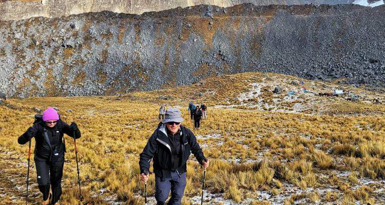 Trekkers push uphill across golden grassland below a sheer rocky escarpment.
