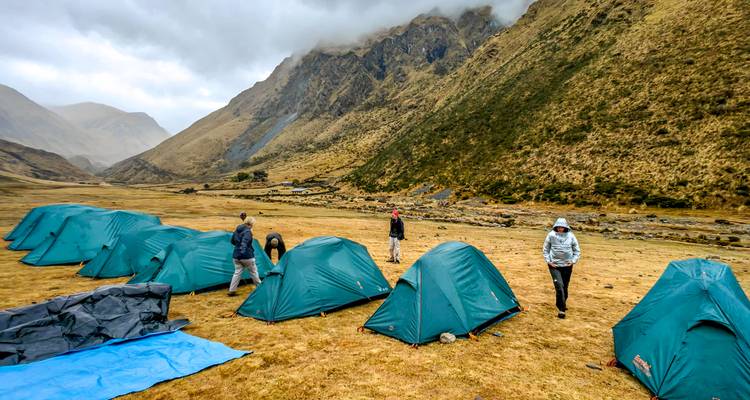 A line of green tents dot a broad misty valley as trekkers walk about camp.