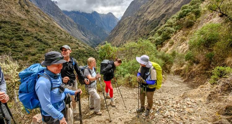 Trekkers pause on a narrow Andean trail surrounded by lush shrubs and deep valleys.