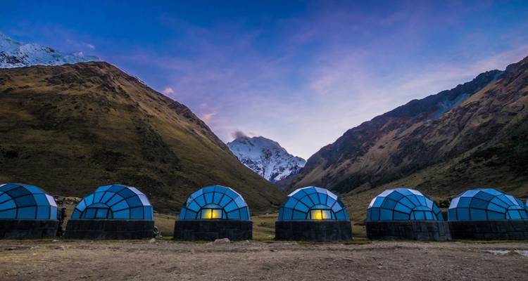 Fila de cúpulas celestes con techo de cristal brillan cálidamente en un valle alto andino al atardecer con un pico nevado al fondo.