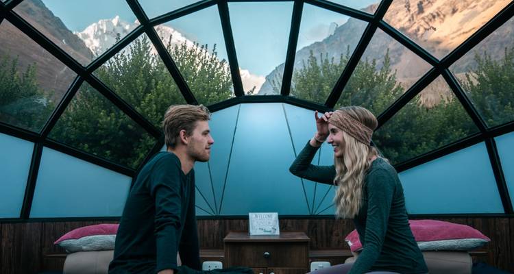 Pareja se relaja dentro de una habitación con domo de cristal con picos nevados visibles a través de la ventana panorámica.