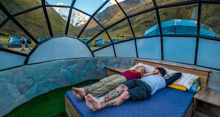 Pareja tatuada descansa en una cama dentro de una cúpula de paredes de piedra con otras cápsulas visibles afuera.