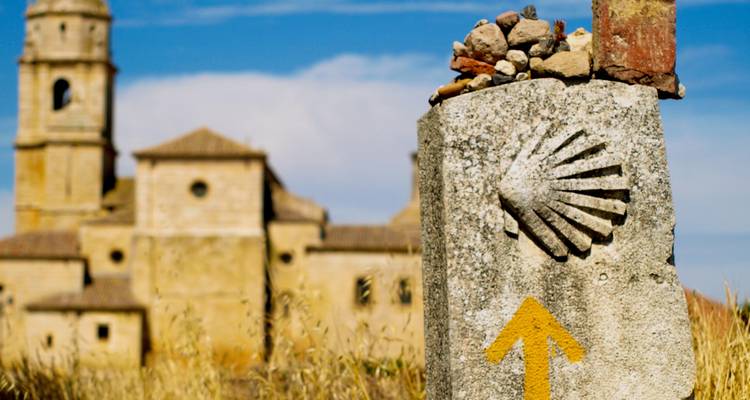 Pilar de piedra con el símbolo de una concha y una flecha amarilla delante de un edificio histórico.