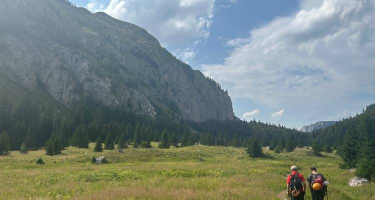 Zwei Rucksacktouristen überqueren eine offene Bergwiese in Richtung steiler bewaldeter Klippen unter fleckigen Wolken.