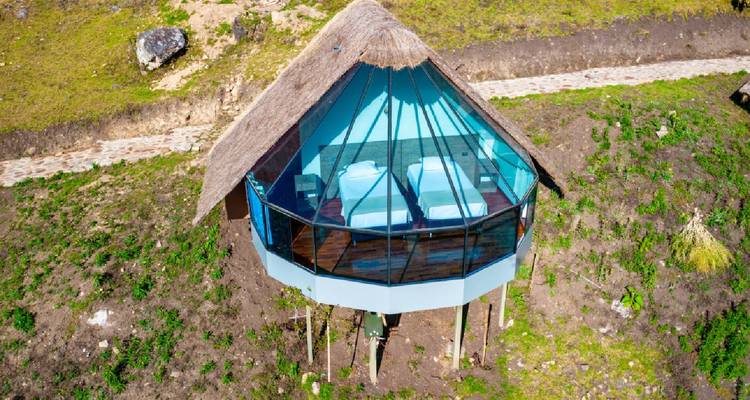 Vista aérea de una cabaña con paredes de cristal y techo de paja sobre pilotes con vista a una ladera montañosa verde.