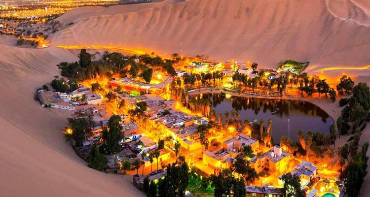 Pueblo oasis resplandeciendo con luces rodeado de dunas de arena por la noche.