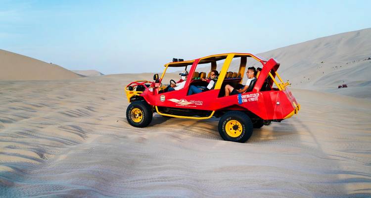 Buggy rojo de dunas con personas sentadas en dunas de arena bajo un cielo azul.