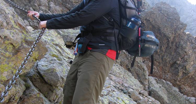 Primer plano de un excursionista agarrado a una cadena en un sendero rocoso.