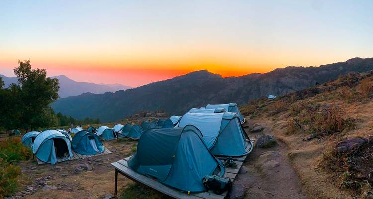 Fila de tiendas azules en un campamento de montaña brillando bajo un cielo naranja al amanecer