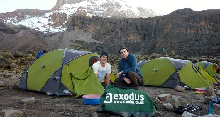 Zwei Menschen posieren neben Zelten mit einem verschneiten Berg im Hintergrund.