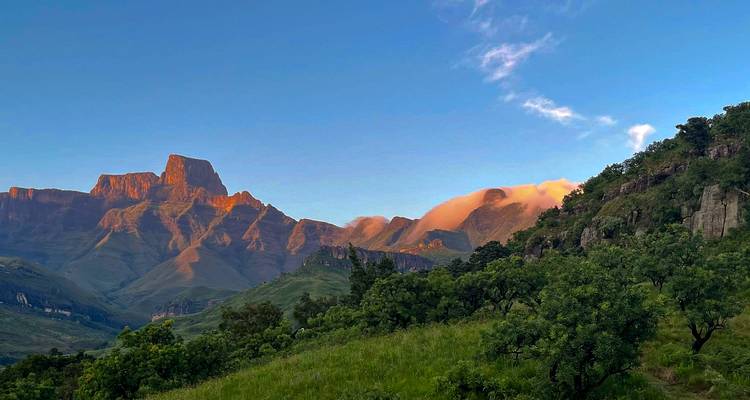 Zonsopganglicht schildert de Drakensberg-pieken oranje boven groene voethellingen en drijvende wolken.