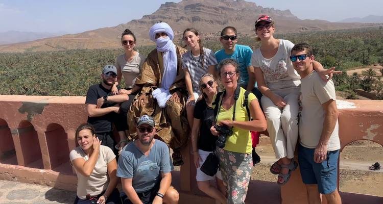 Groupe de voyageurs posant avec un guide local en tenue traditionnelle surplombant une vallée d'oasis luxuriante et des montagnes.