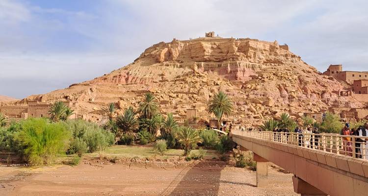 Kasbah historique en terre d'Aït Benhaddou s'élevant au-dessus d'un lit de rivière à sec avec pont et palmiers.