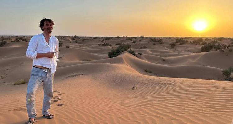Voyageur solitaire debout sur les dunes dorées au coucher du soleil tenant des lunettes de soleil au milieu du paysage ondulant du Sahara.