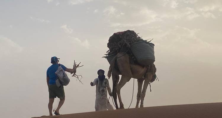 Voyageur remettant du bois de chauffage ramassé à un guide à côté d'un chameau chargé sur une crête désertique venteuse.