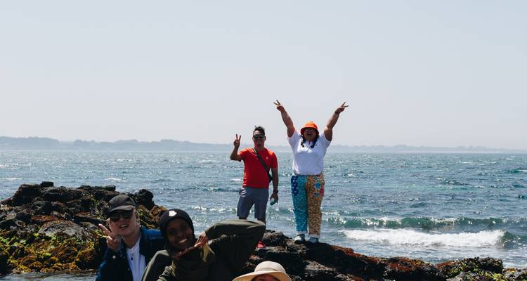 Viajeros posando con signos de paz en la orilla rocosa con mar brillante y olas suaves