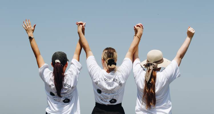 Tres mujeres vistas desde atrás levantan las manos unidas triunfalmente contra un cielo azul claro
