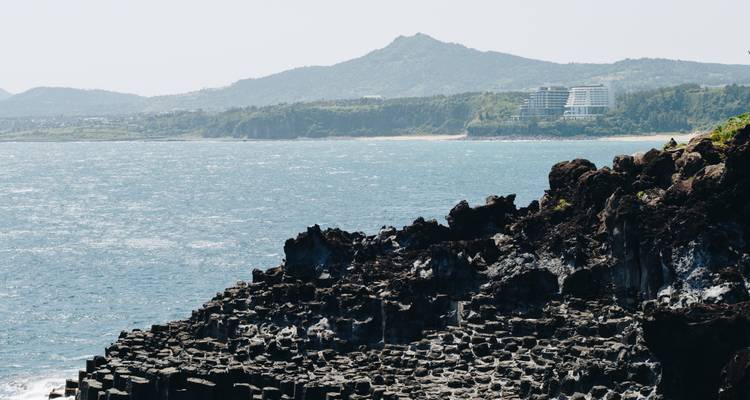 Formaciones rocosas de acantilados de columnas basálticas escarpadas a lo largo de la costa ventosa de Jeju con mar azul y colinas distantes