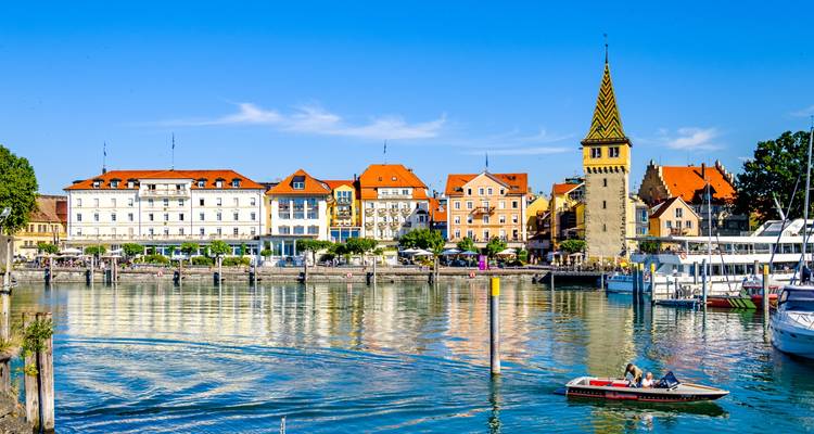 Helle und lebendige Uferpromenade mit Booten und farbenfrohen Gebäuden, die sich im Wasser spiegeln.