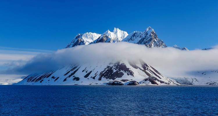 Majestueuze met sneeuw bedekte bergen gedeeltelijk bedekt door wolken boven blauw water.