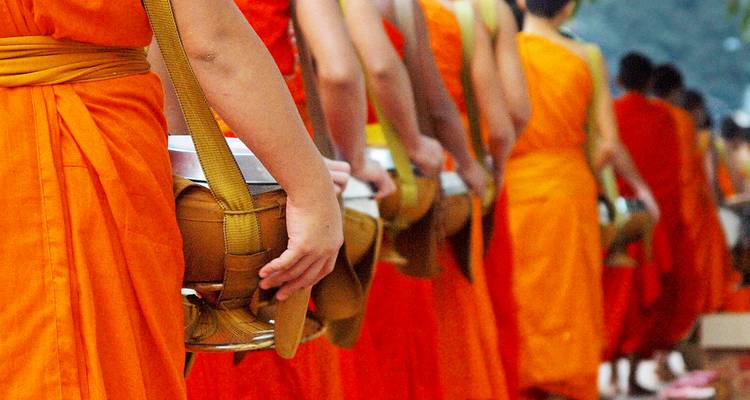 Line of Buddhist monks in bright orange robes carrying alms bowls during morning procession.