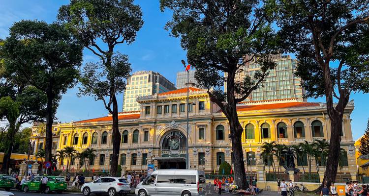 Bureau de poste central de Saigon encadré par de grands arbres et l'animation de rue dans la lumière de fin de journée.