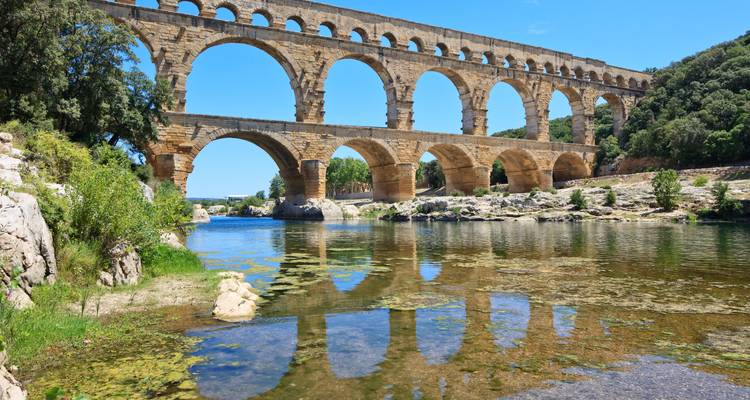 Een historisch stenen aquaduct dat zich boogvormig over een heldere rivier uitstrekt, omgeven door weelderig groen.