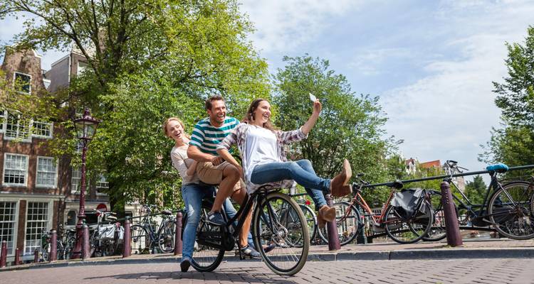 Drei Freunde teilen sich ein Fahrrad, während sie ein verspieltes Selfie auf einer Amsterdamer Brücke machen.