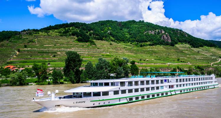 Long white river cruise ship gliding past terraced vineyards and forested hills along the Danube.