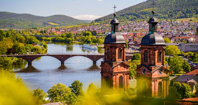 Église baroque à tours jumelles au-dessus d'une rivière sereine enjambée par un élégant pont de pierre, encadrée par des forêts.