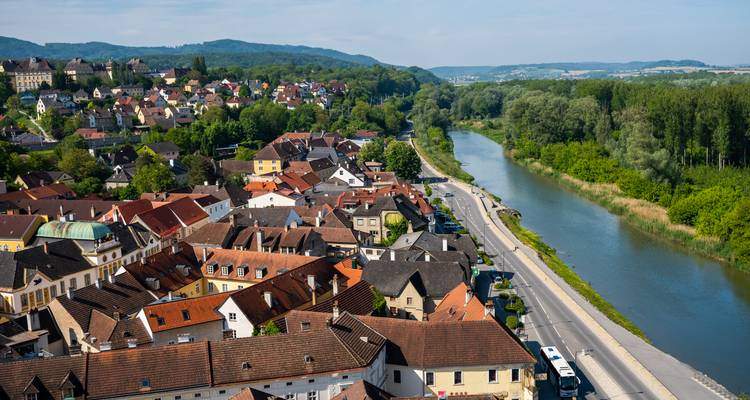 Vista aérea de un pueblo austriaco compacto a orillas del río con hileras de tejados de tejas rojas junto a un río verde tranquilo y colinas boscosas ondulantes.