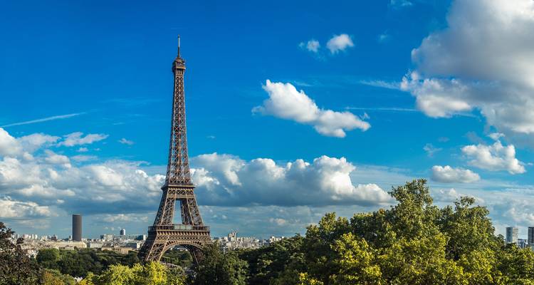 Vista icónica de la Torre Eiffel elevándose sobre árboles frondosos contra un cielo azul vívido salpicado de nubes blancas.