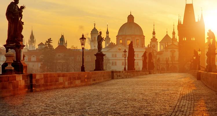 Golden sunrise light streaming through statues along Prague’s Charles Bridge with silhouettes of spires in the mist.