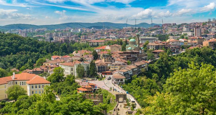 Üppige grüne Hügel umhüllen die Altstadt und Festung von Veliko Tarnovo mit ihren roten Dächern unter blauem Himmel.
